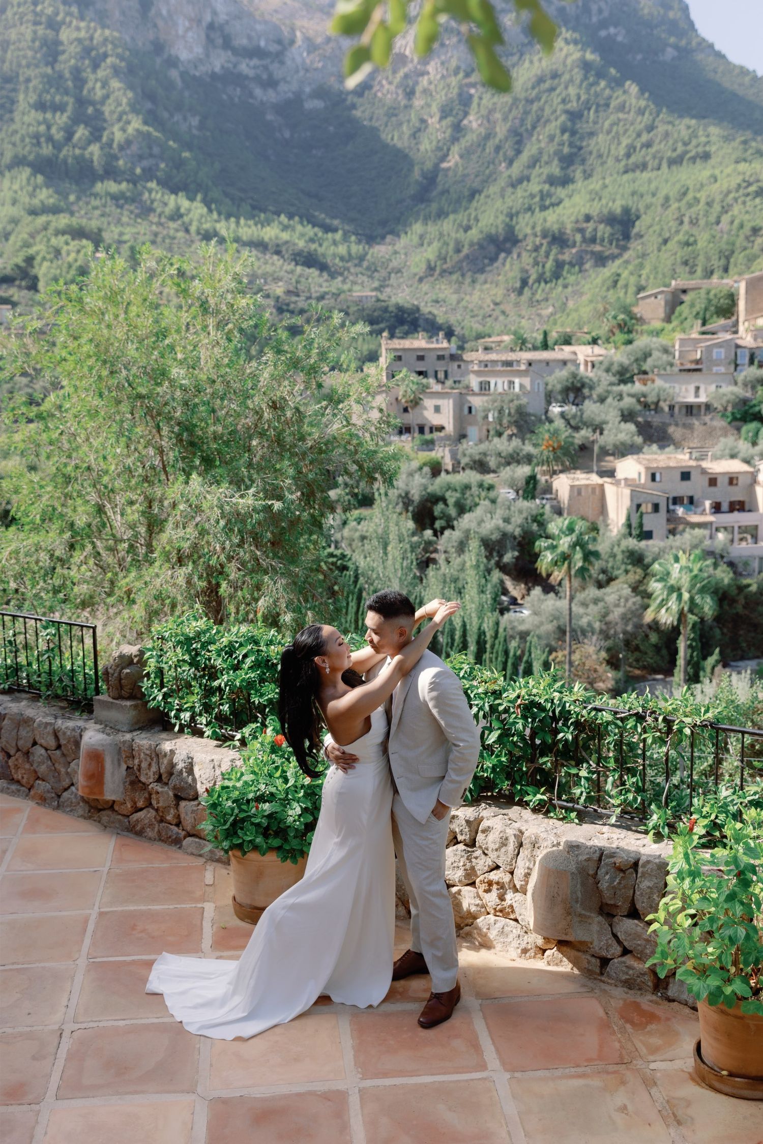 Bride and groom embrace on a sunlit terrace with lush mountain village backdrop in Mallorca, Spain.