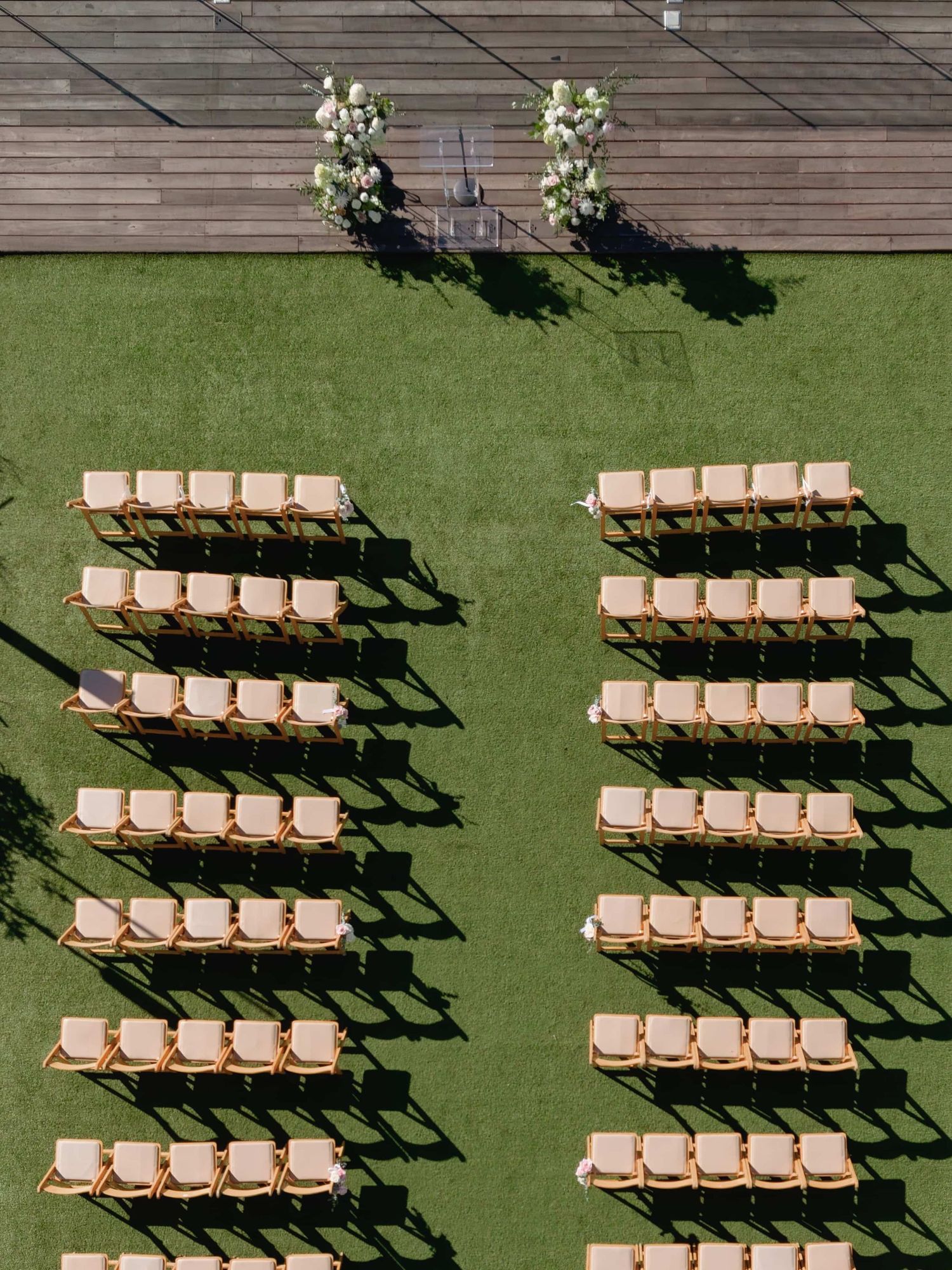 Aerial view of outdoor wedding ceremony setup with rows of wooden chairs on green grass and floral arch.