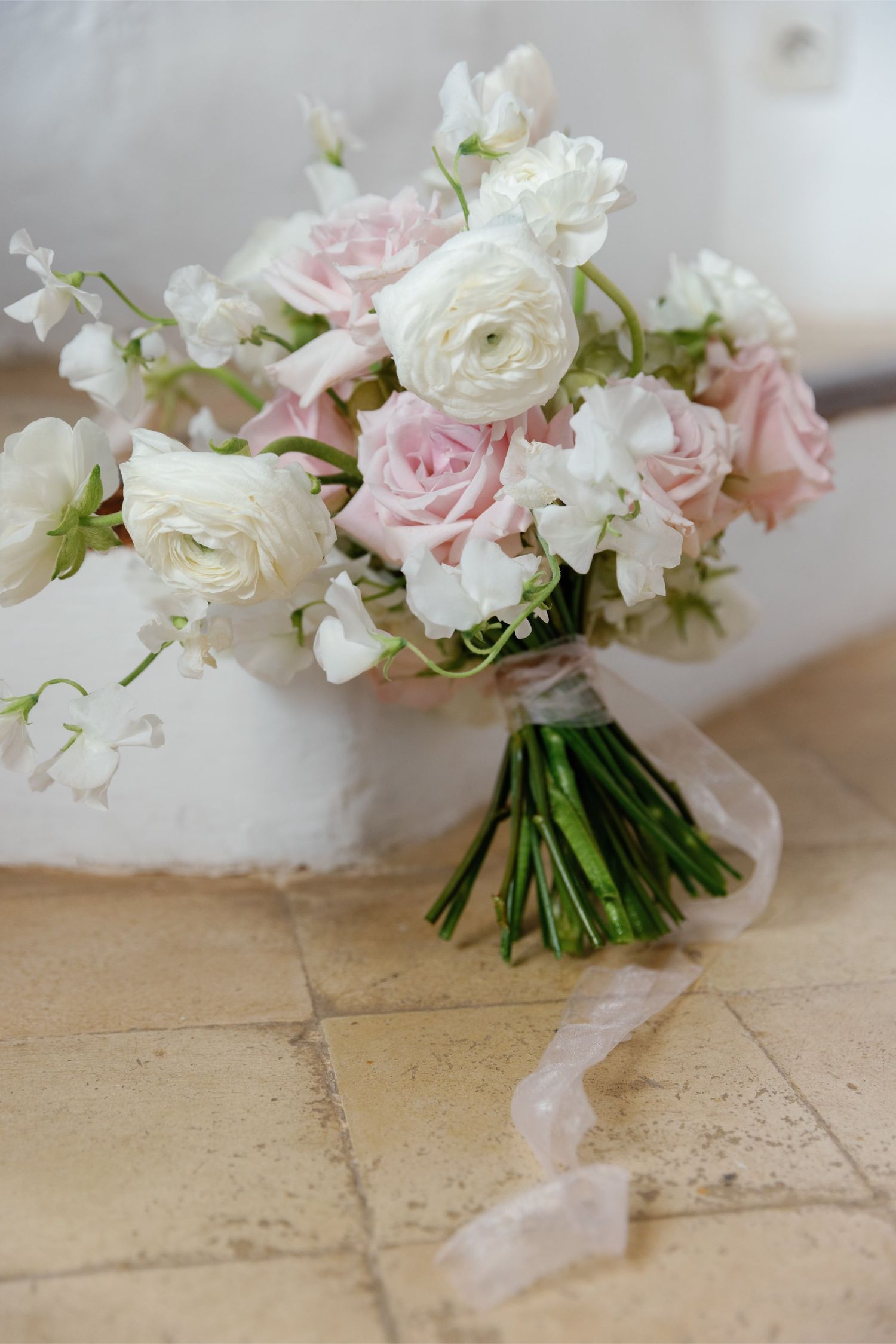 Elegant bridal bouquet of white ranunculus, blush pink roses, and sweet peas tied with ribbon on stone floor.