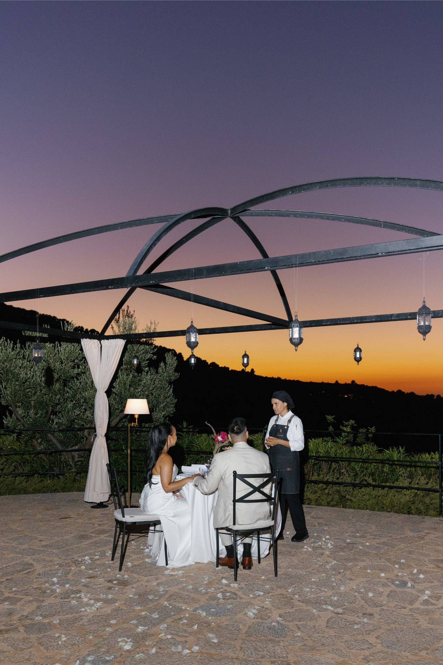 Couple dining outdoors under elegant gazebo with hanging lanterns at sunset, served by a waiter.