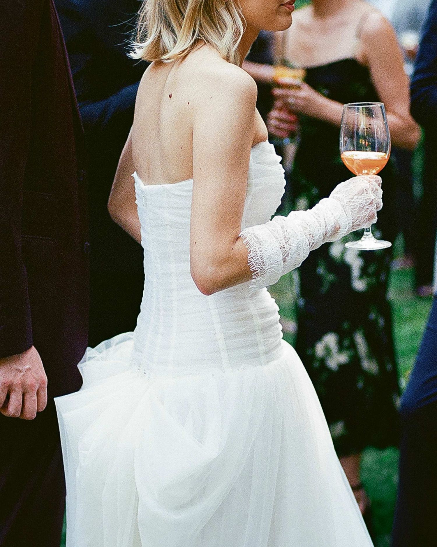Bride in strapless white wedding gown holding rose wine glass, wearing lace gloves at outdoor wedding reception.