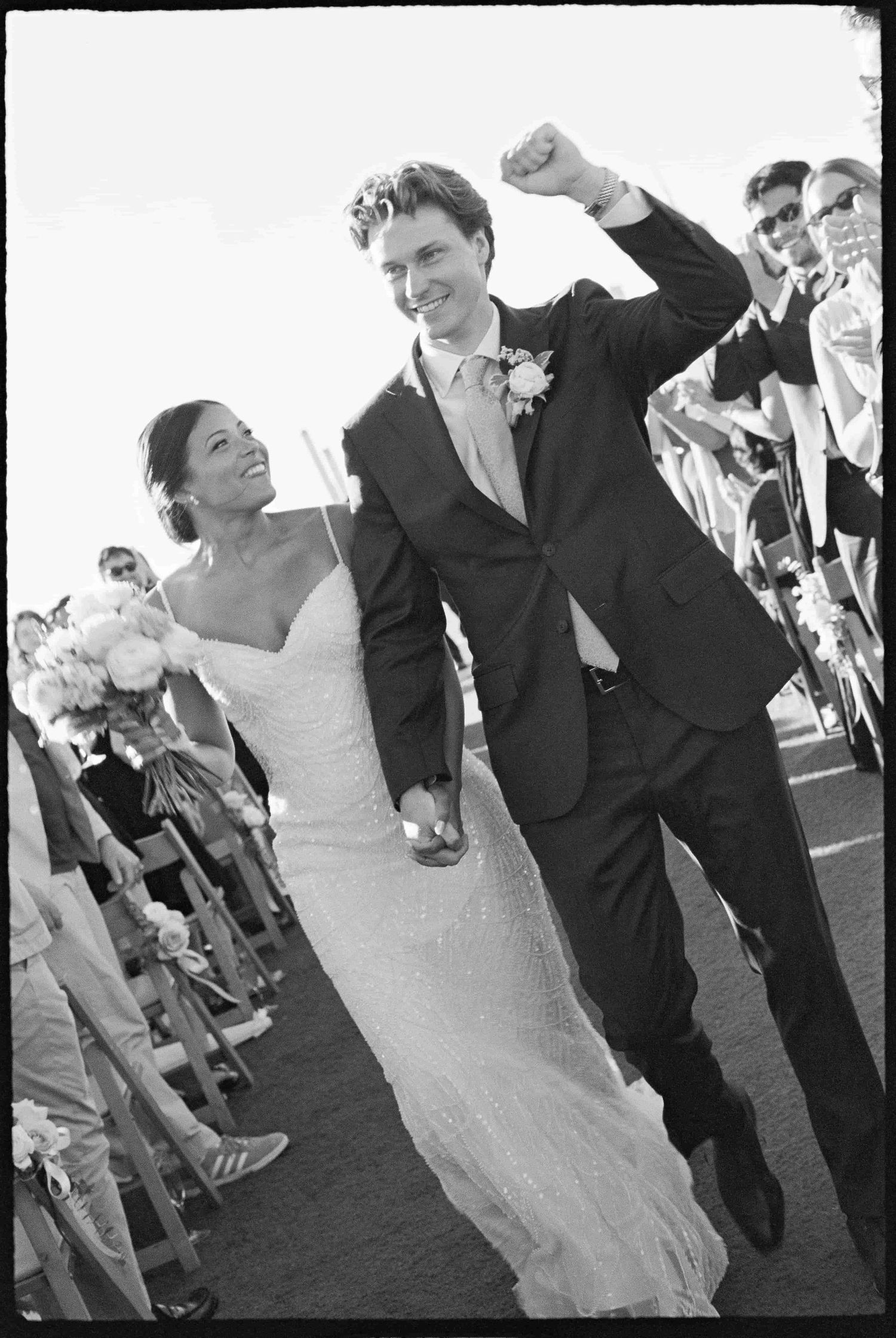 Black and white photo of a joyful newlywed couple celebrating after their wedding ceremony with cheering guests lining the aisle.