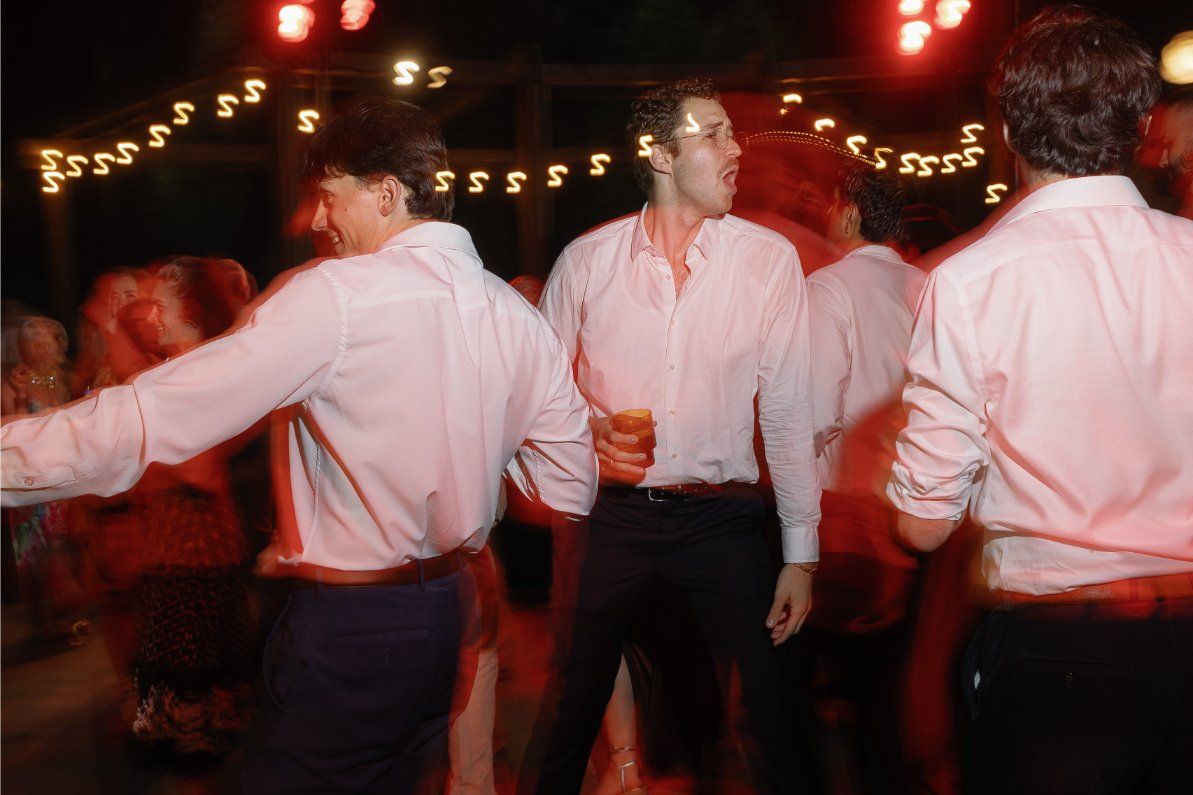 Party guests in white shirts dance under red lighting and string lights at a nighttime celebration.