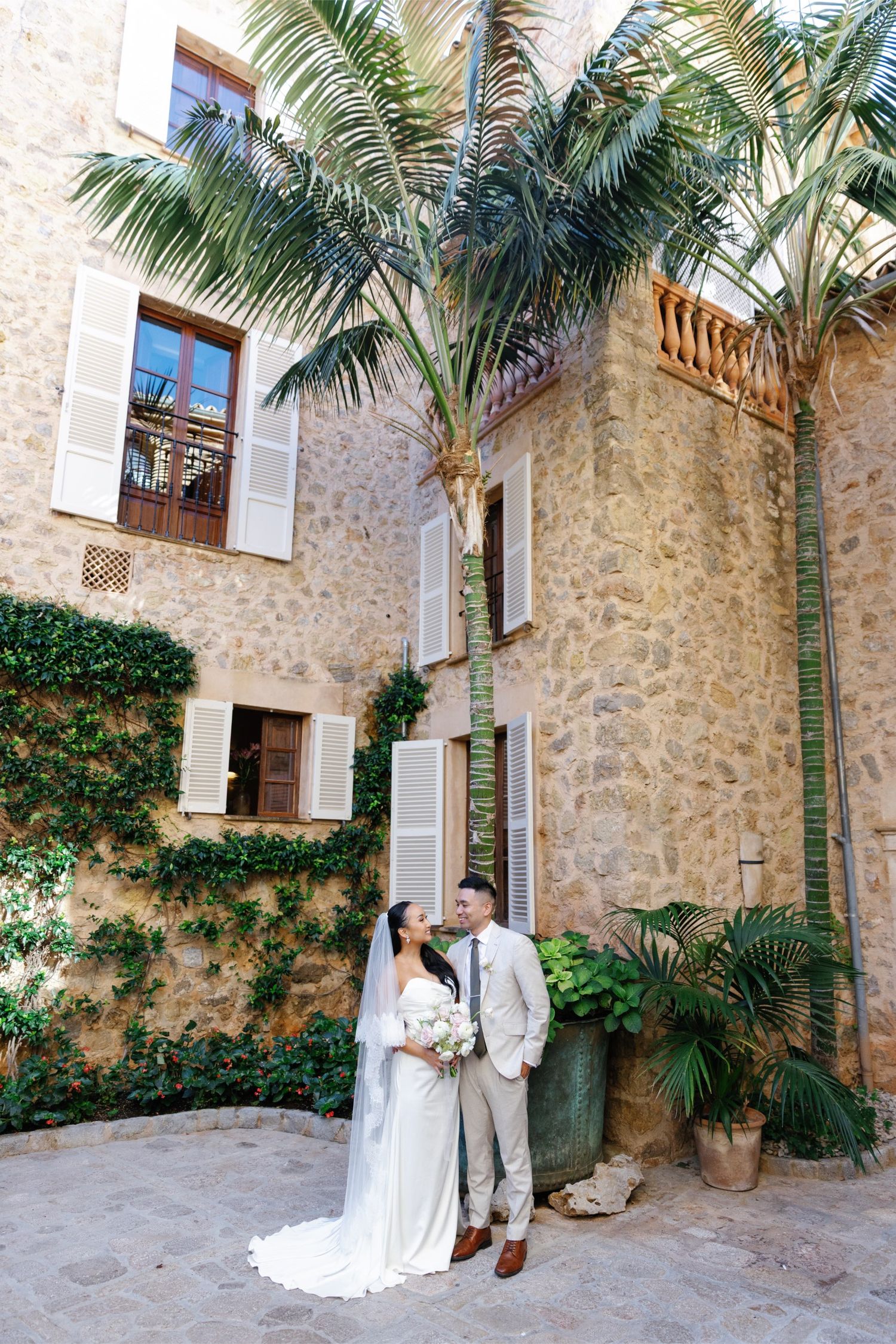 Bride and groom pose in a rustic stone courtyard with palm trees and ivy-covered walls at a destination wedding.
