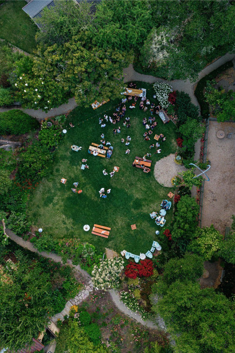Aerial view of an outdoor garden party with scattered picnic tables and people gathered on a lush green lawn surrounded by paths.