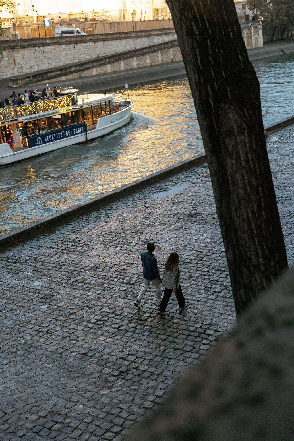 Two people walk along cobblestone riverbank at sunset as tourist boat passes on Seine River in Paris, France.