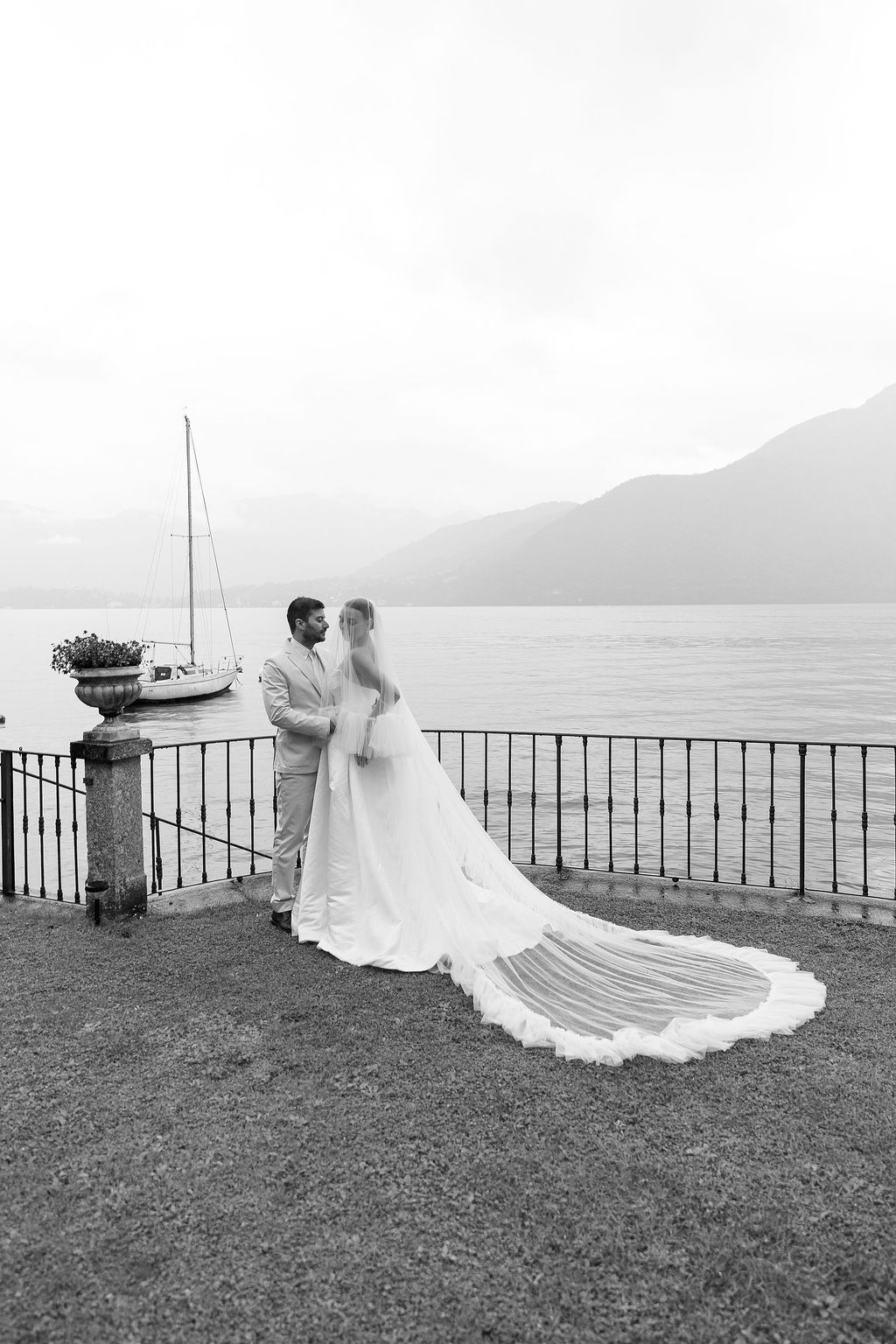 A romantic black and white wedding photo series showing a couple by a lakeside railing with mountains in the distance in Lake Como.