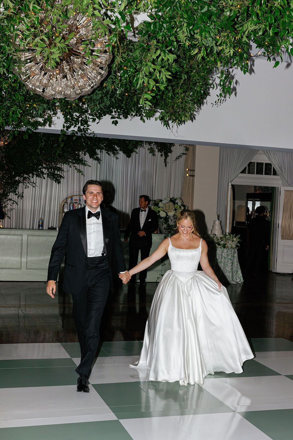 A couple in formal wedding attire walk together on a black and white checkered dance floor at a reception venue.
