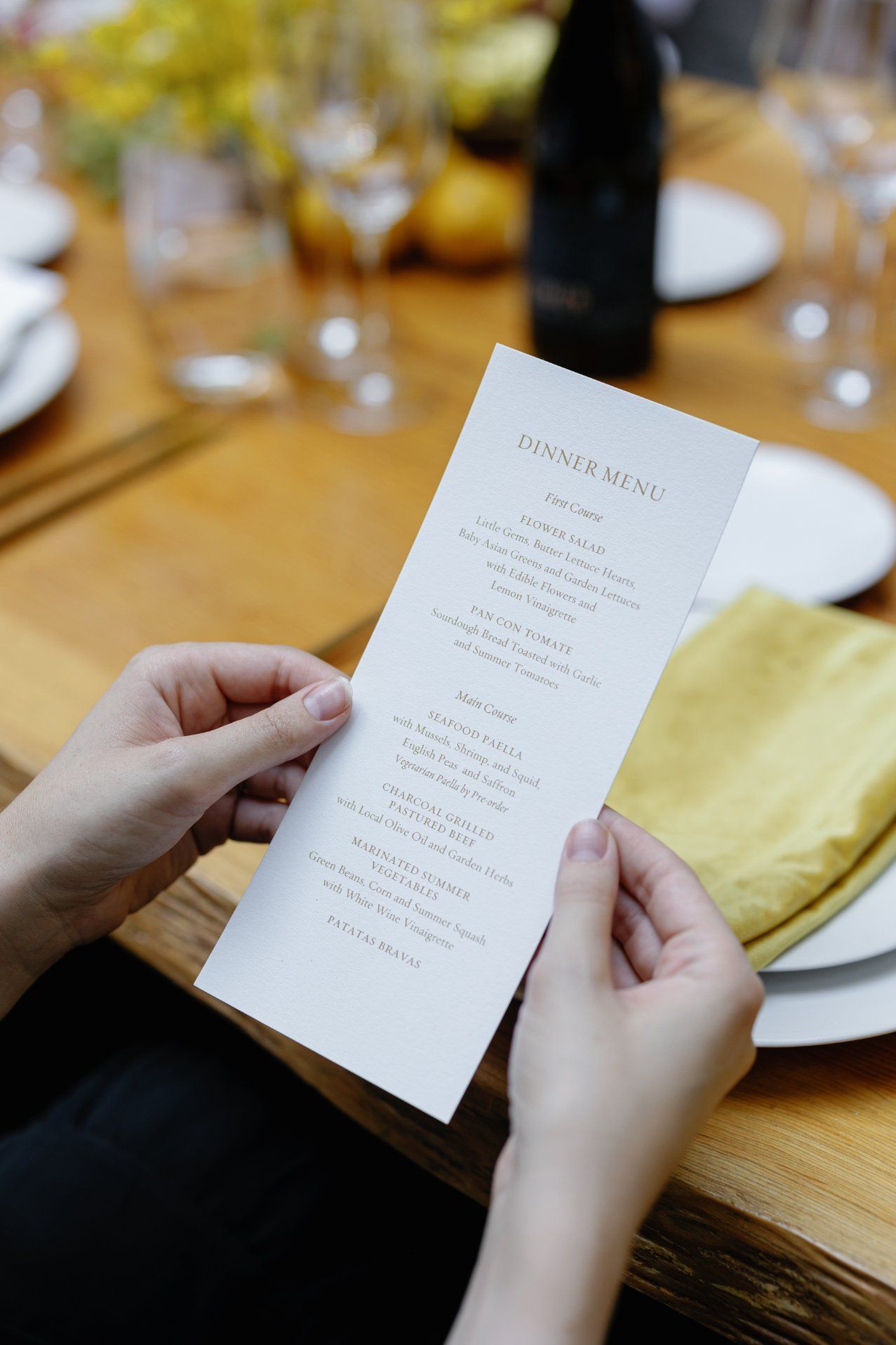 Hands holding an elegant dinner menu at a formal table setting with wine glasses and yellow napkins.