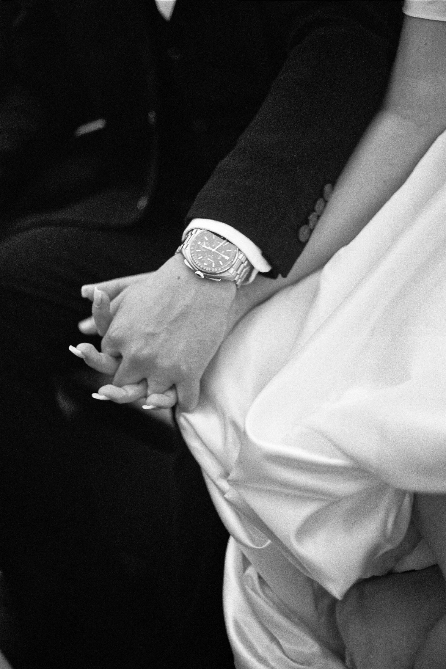 Black and white close-up of hands holding together, with a dress watch visible on one wrist against formal attire.
