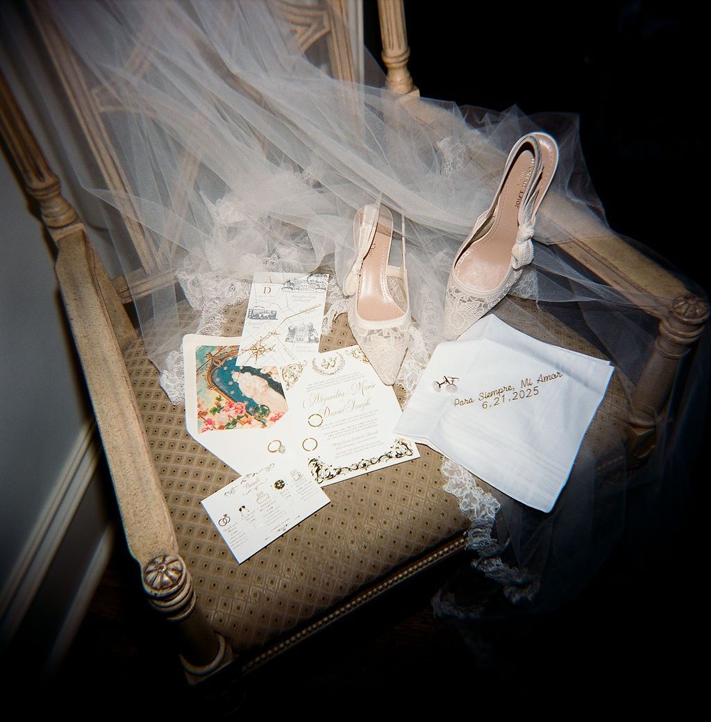Elegant beige bridal shoes sit with scattered wedding invitation cards and delicate tulle veil on antique wooden chair.