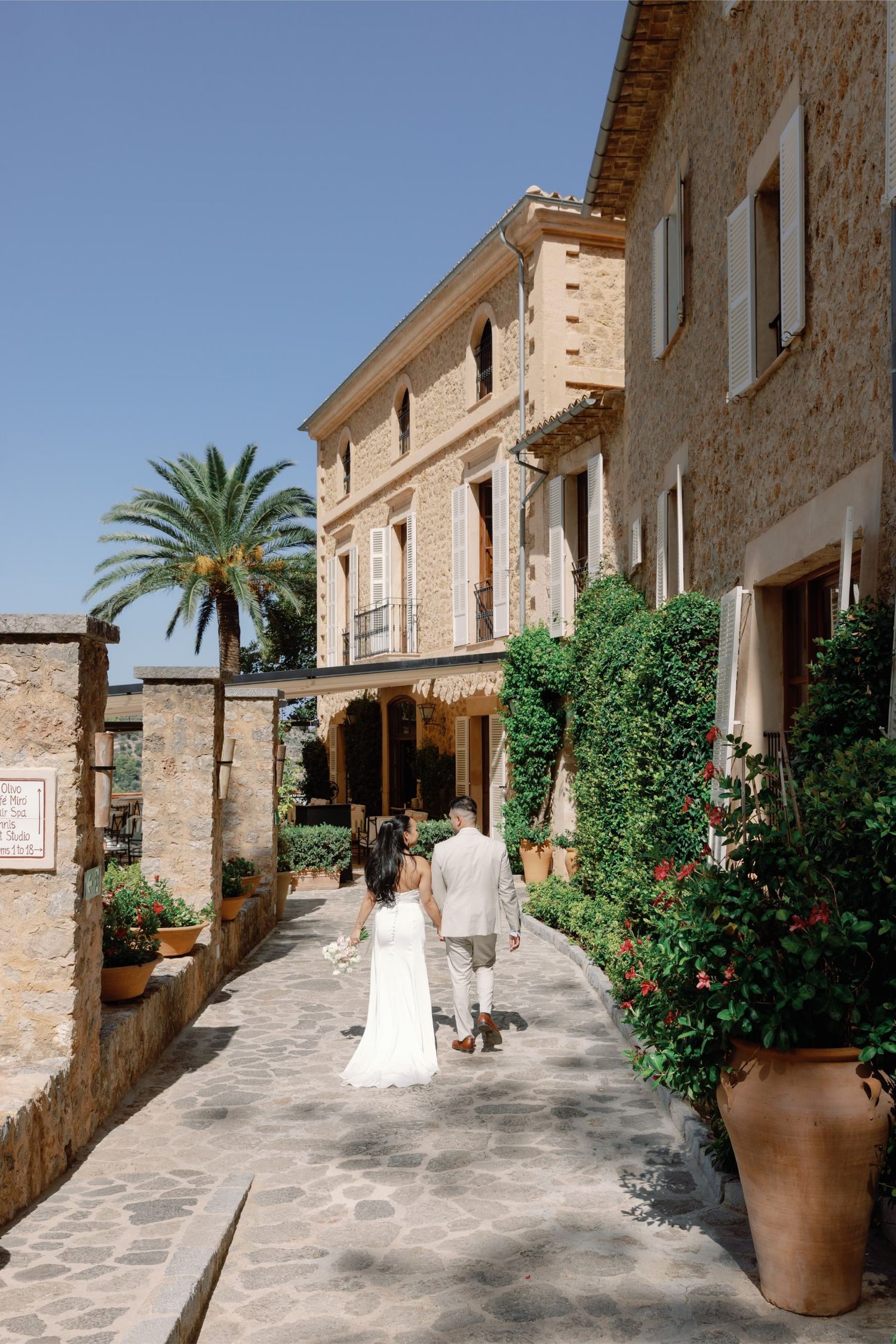 Couple walking hand in hand through a charming stone-paved Mediterranean village street lined with lush greenery.