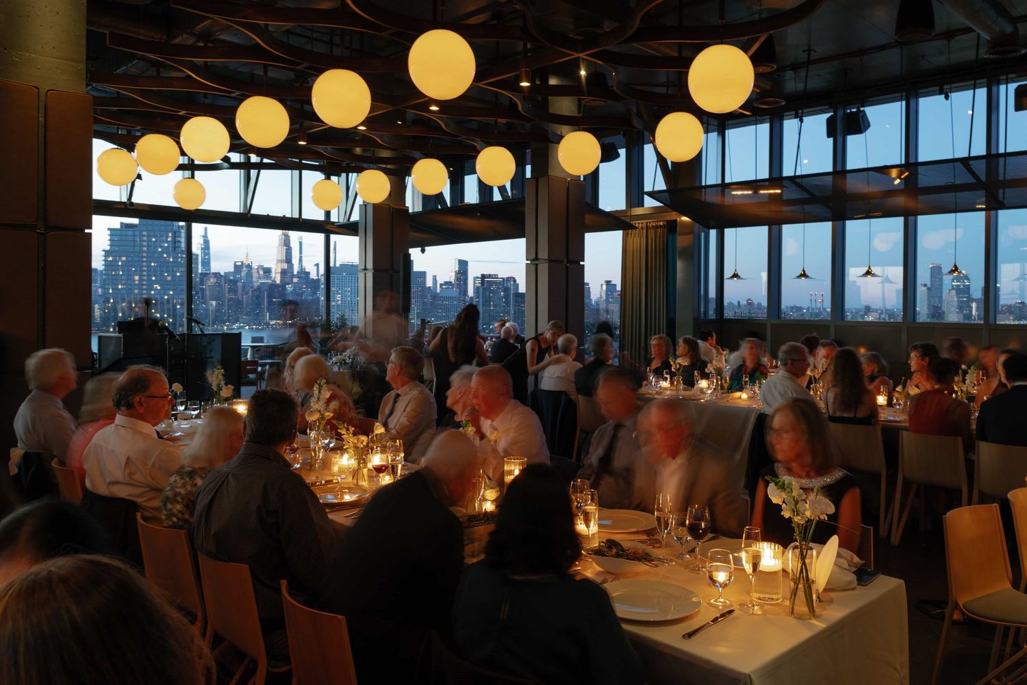 Elegant dinner event in a NYC restaurant with round globe lights, candlelit tables, and a stunning city skyline view.
