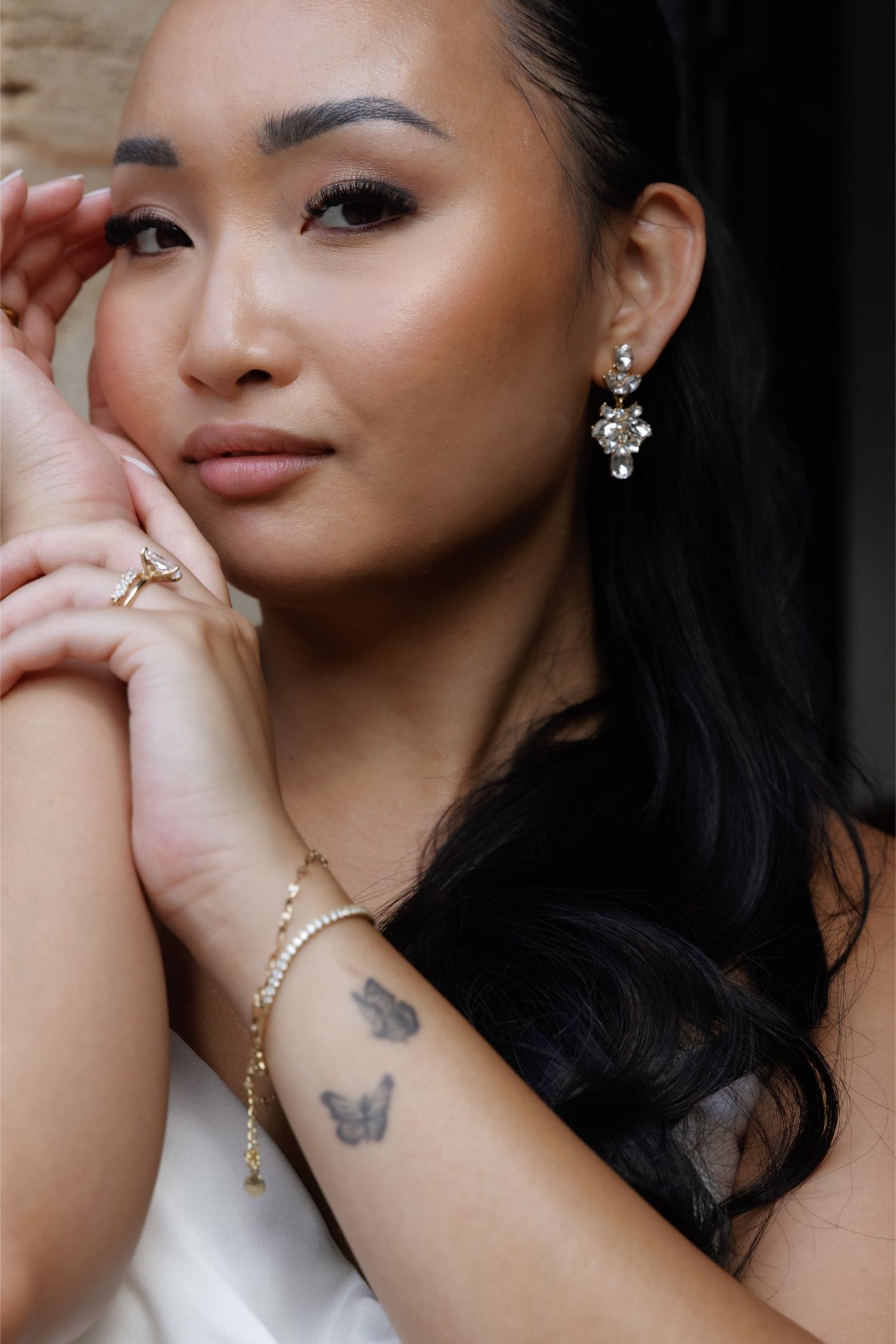 Woman with butterfly tattoo and pearl bracelet wearing crystal drop earrings, posing thoughtfully with hands near face.
