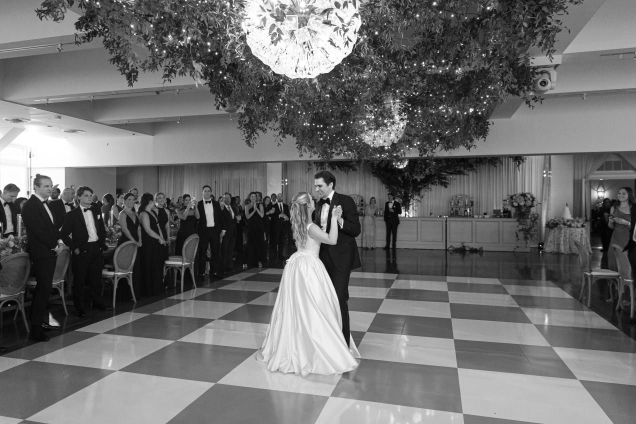 Couple shares their first dance on a black and white checkered floor at an elegant indoor wedding reception.