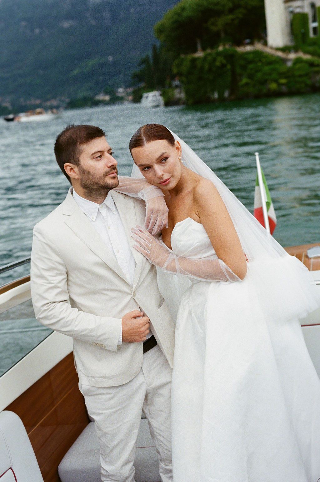 Newlyweds in white attire embrace on wooden boat against Lake Como backdrop.