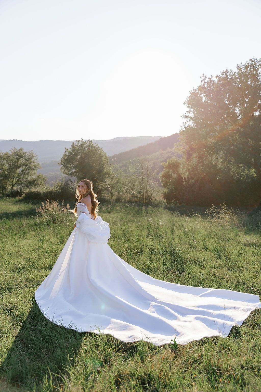 A flowing white dress train spreads across green grass with mountains in the background in Tuscany, Italy.