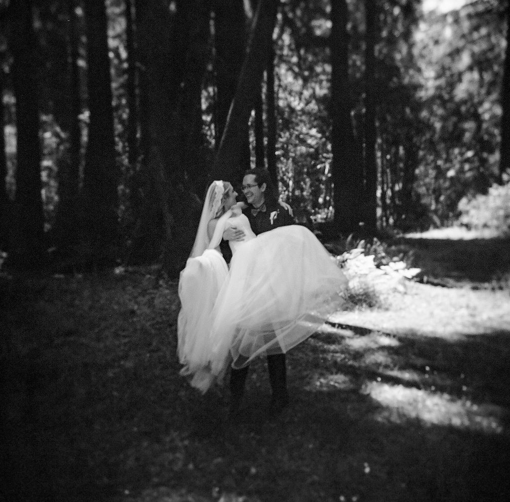 A romantic black and white wedding photo in a forest setting with sunlight filtering through the trees.
