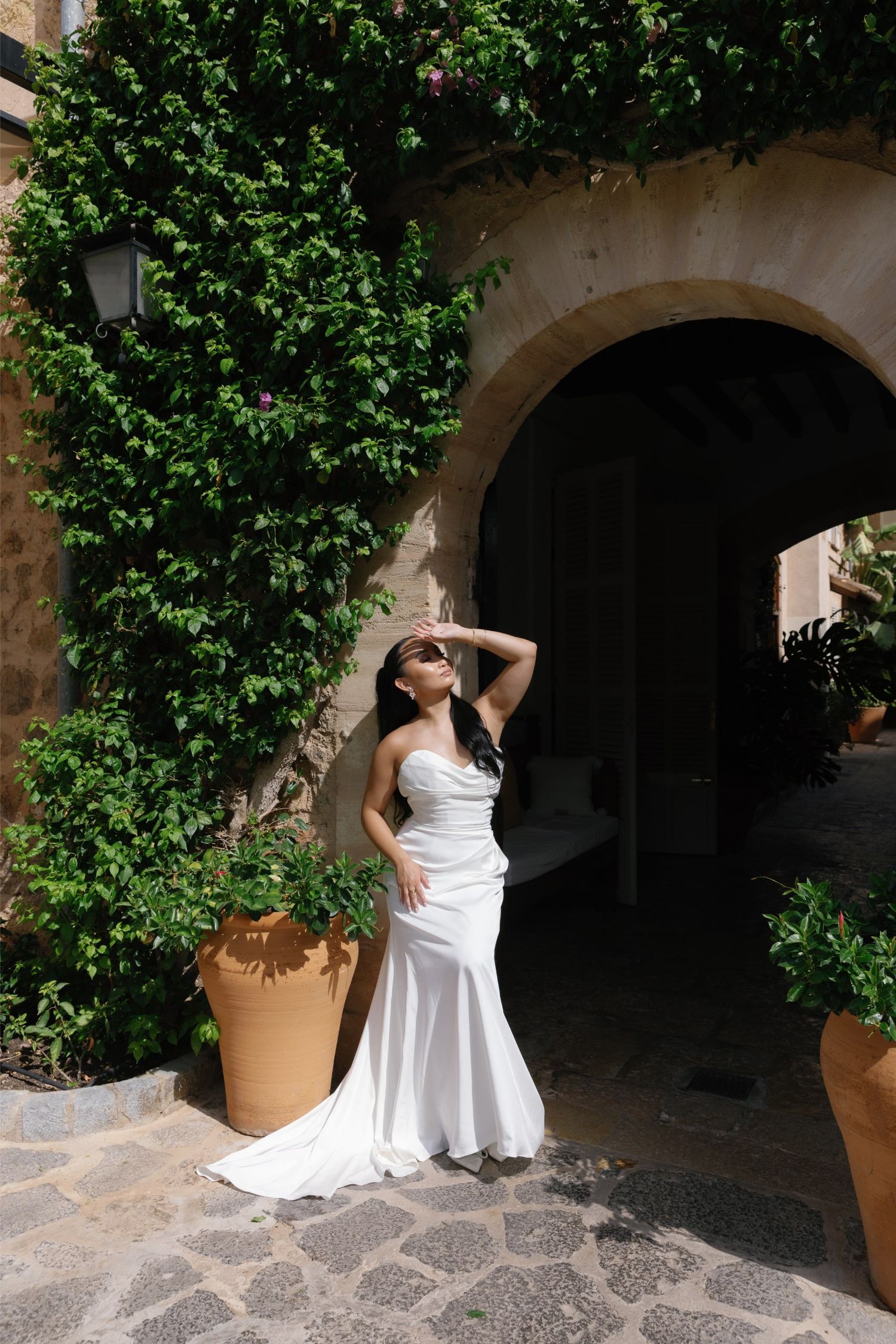 Bride in white strapless gown poses by stone archway with lush green ivy, terracotta pots on cobblestone patio.