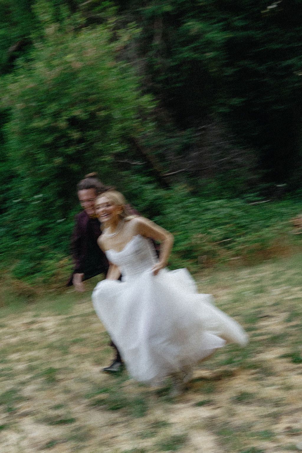 Motion blur captures running figures in white dress through a green wooded area.