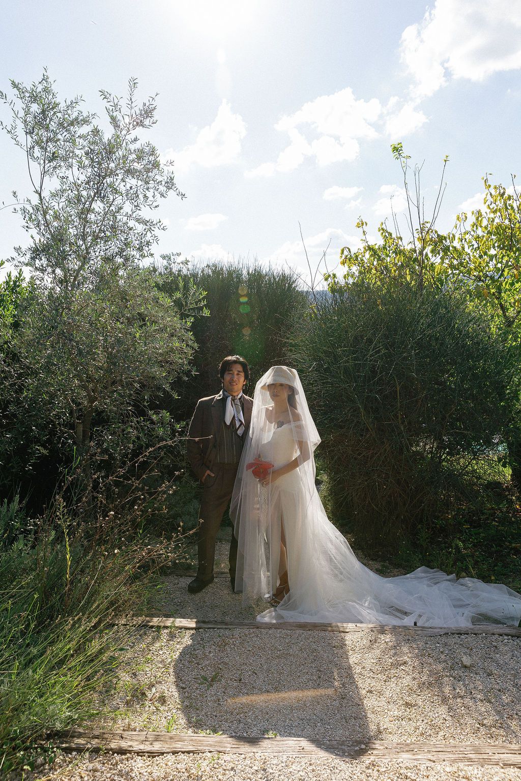 A wedding couple poses among olive trees on a sunlit gravel path in Tuscany, Italy.