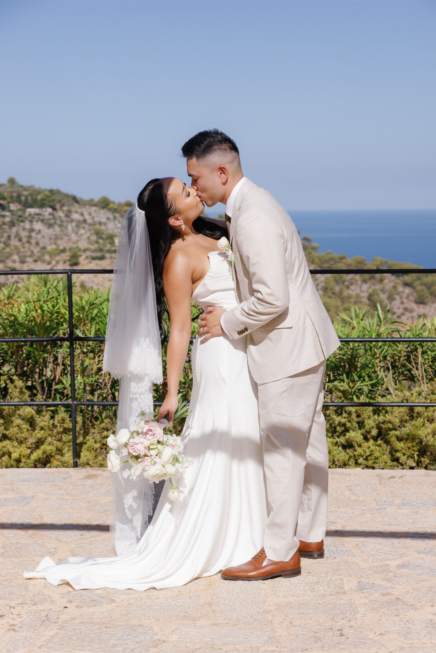 Bride and groom sharing a kiss on a coastal terrace, bride holding a bouquet in a white gown.