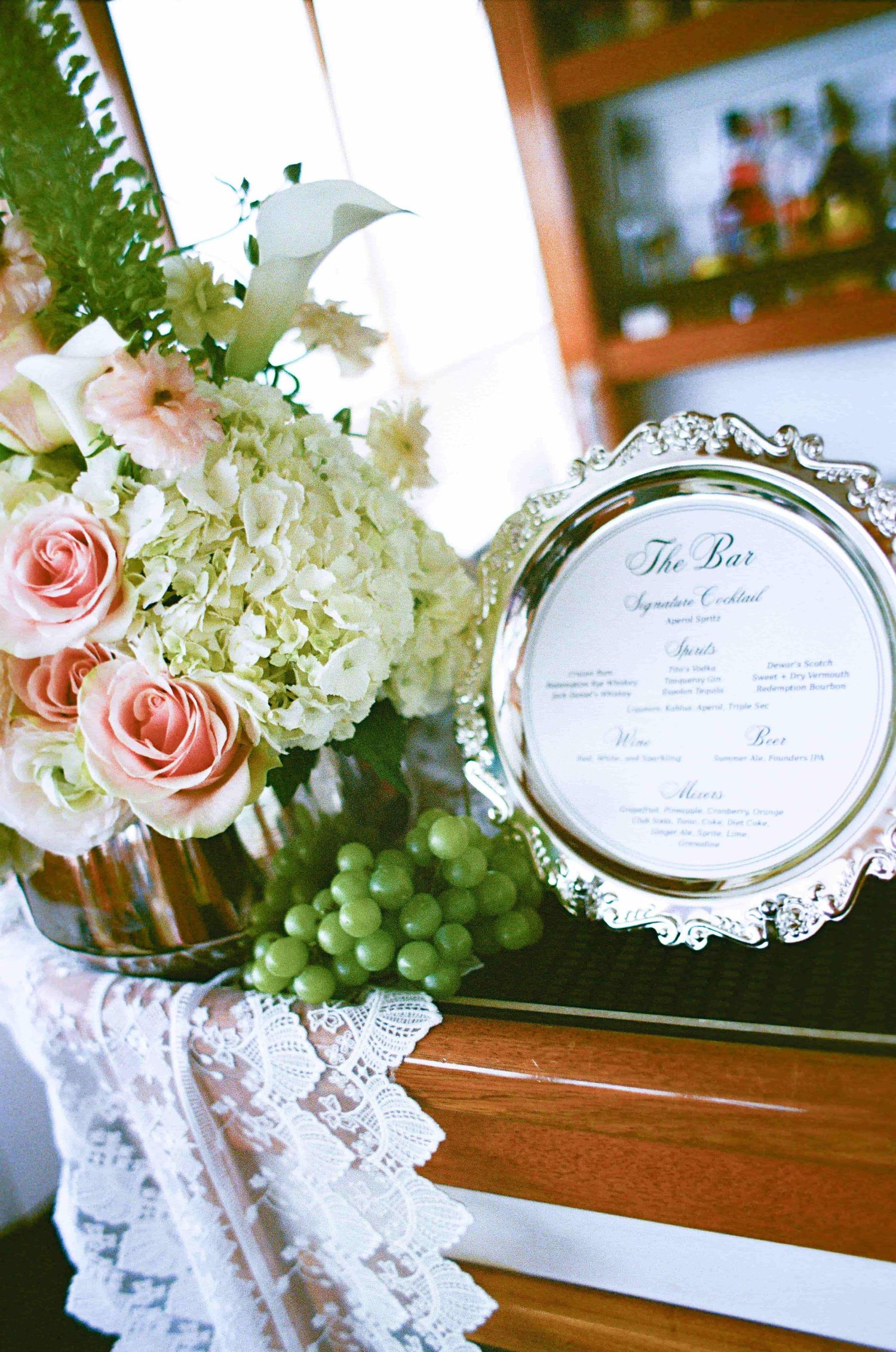 Elegant wedding bar menu in silver frame beside pink roses, white hydrangeas, and green grapes on lace-draped table.