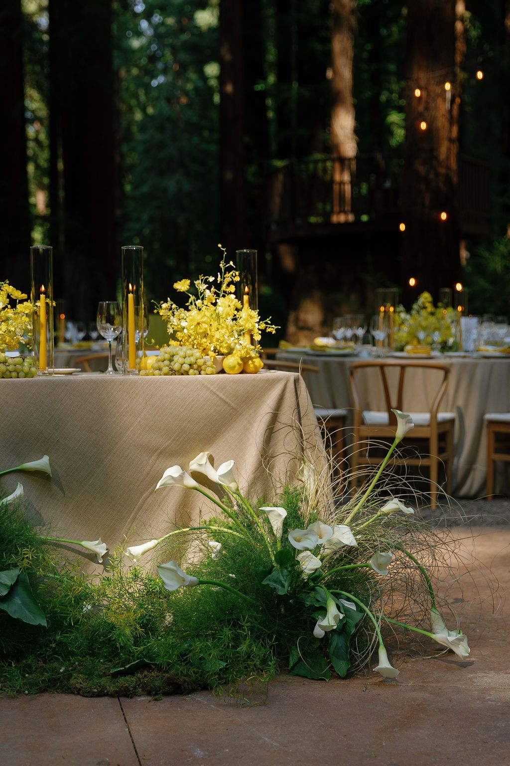 Elegant outdoor wedding reception table with yellow floral arrangements and greenery in a forest setting.