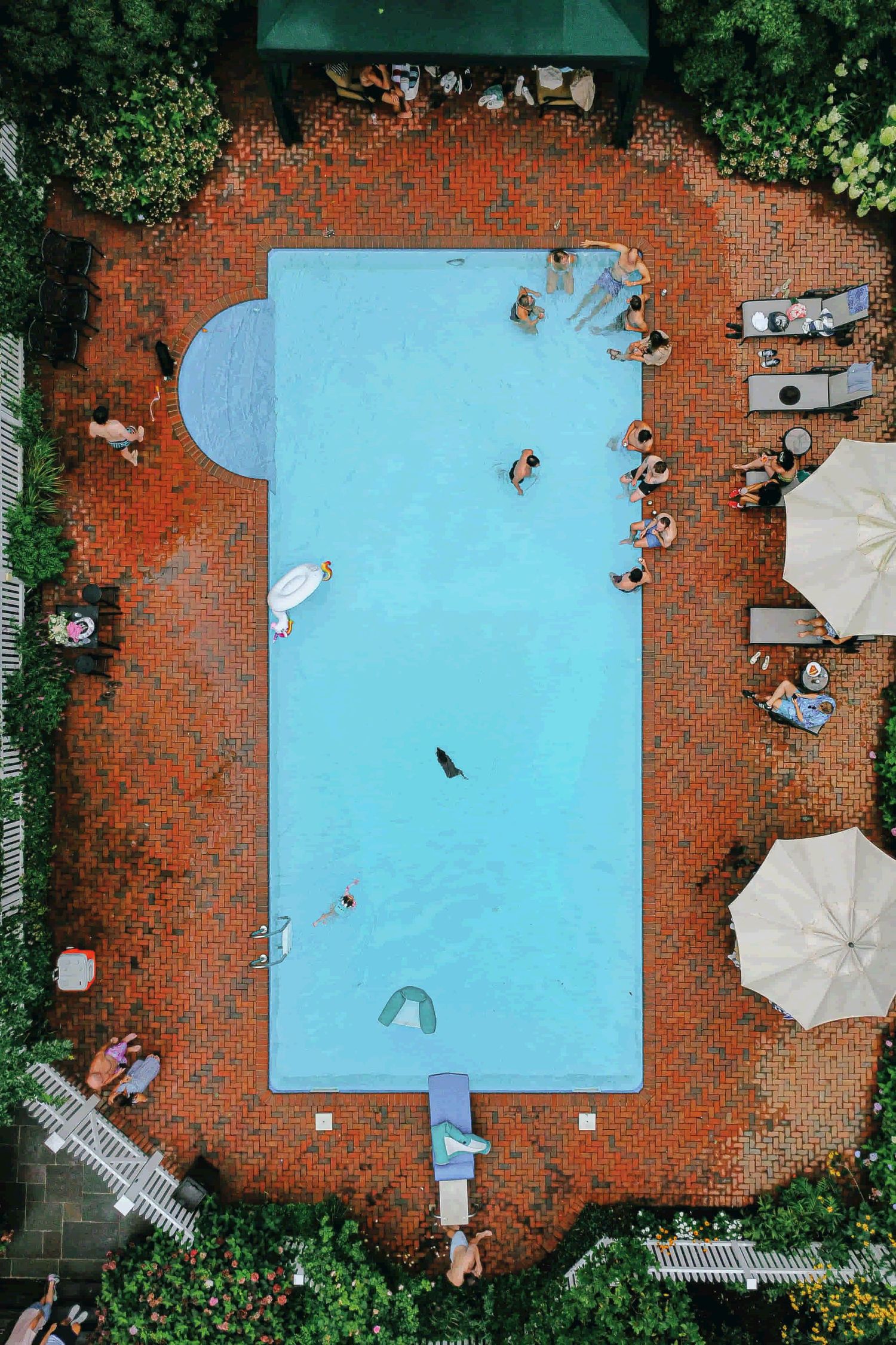 Aerial view of a rectangular swimming pool with turquoise water surrounded by red brick decking and white umbrellas.