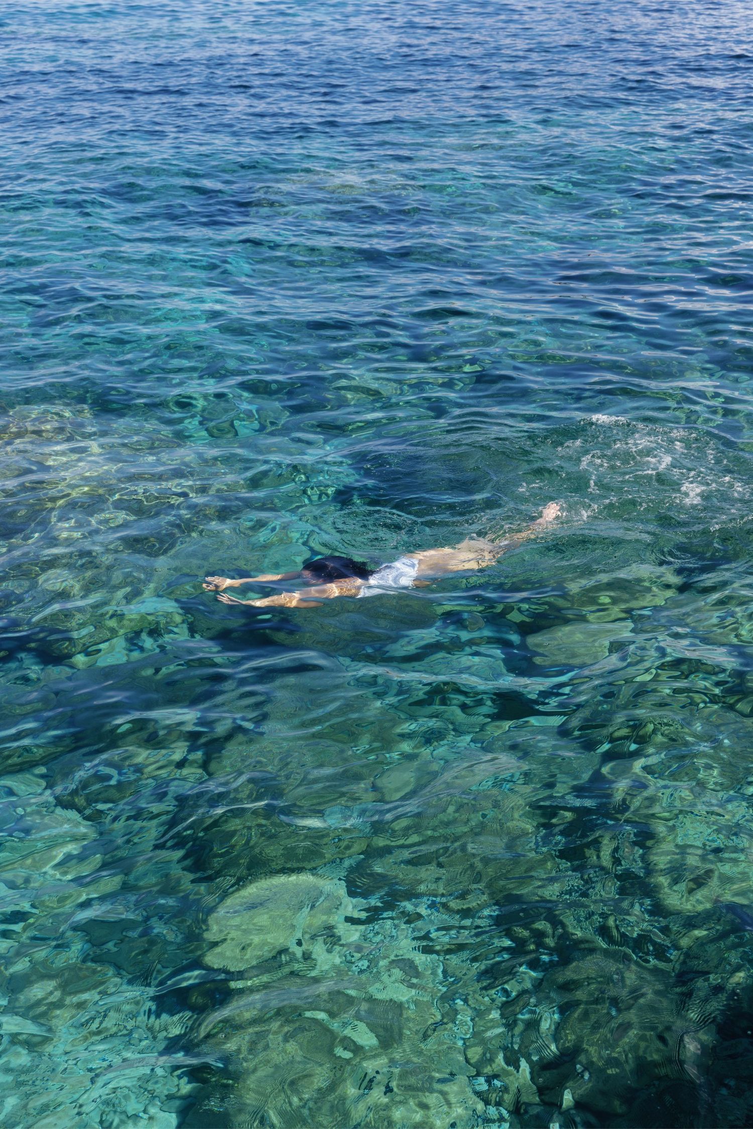 Person snorkeling in clear turquoise ocean water above rocky reef visible through the transparent sea.