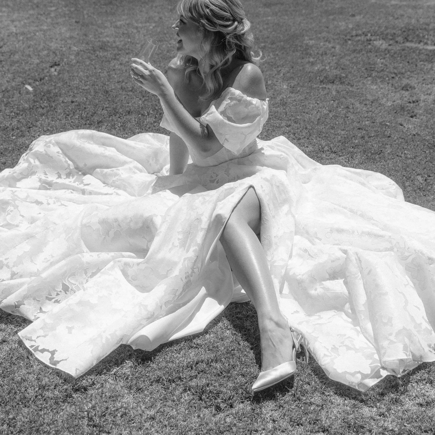 Bride in flowing white dress sitting in grass showing ballet flats in black and white photography series.
