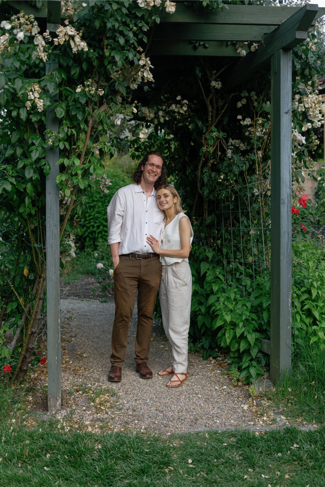 Two figures share a quiet moment under a rustic pergola covered with climbing vines in a garden setting.