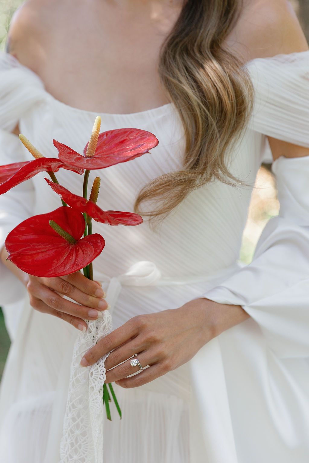 A person in a white dress holds a vibrant red anthurium flower against a light background in Tuscany, Italy.