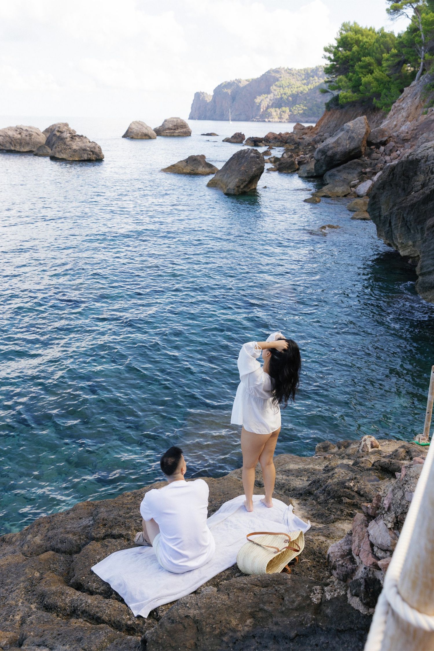 Two women relaxing on rocky coastal cliffs beside crystal blue Mediterranean waters with scenic rock formations in background.