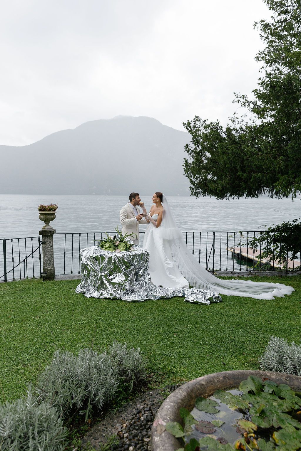 A romantic wedding couple sits on a stone wall overlooking a misty lake and mountains in an elegant garden setting in Lake Como.