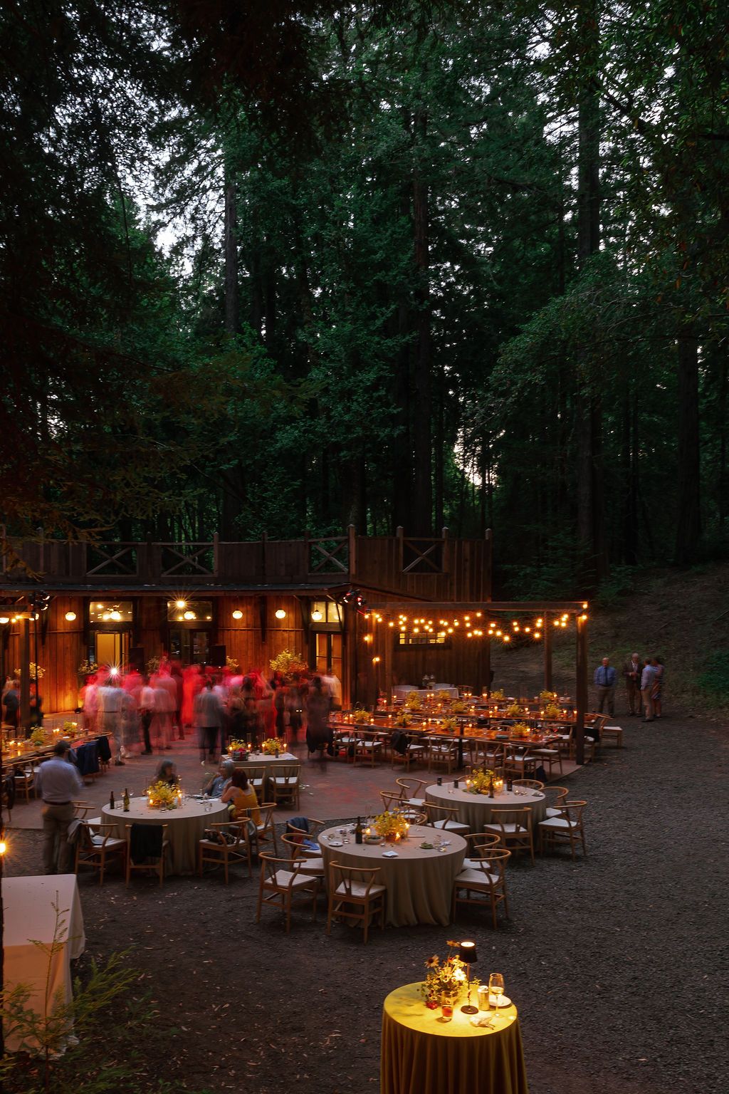 Outdoor evening wedding reception in a forest with tables illuminated by warm string lights and candles.