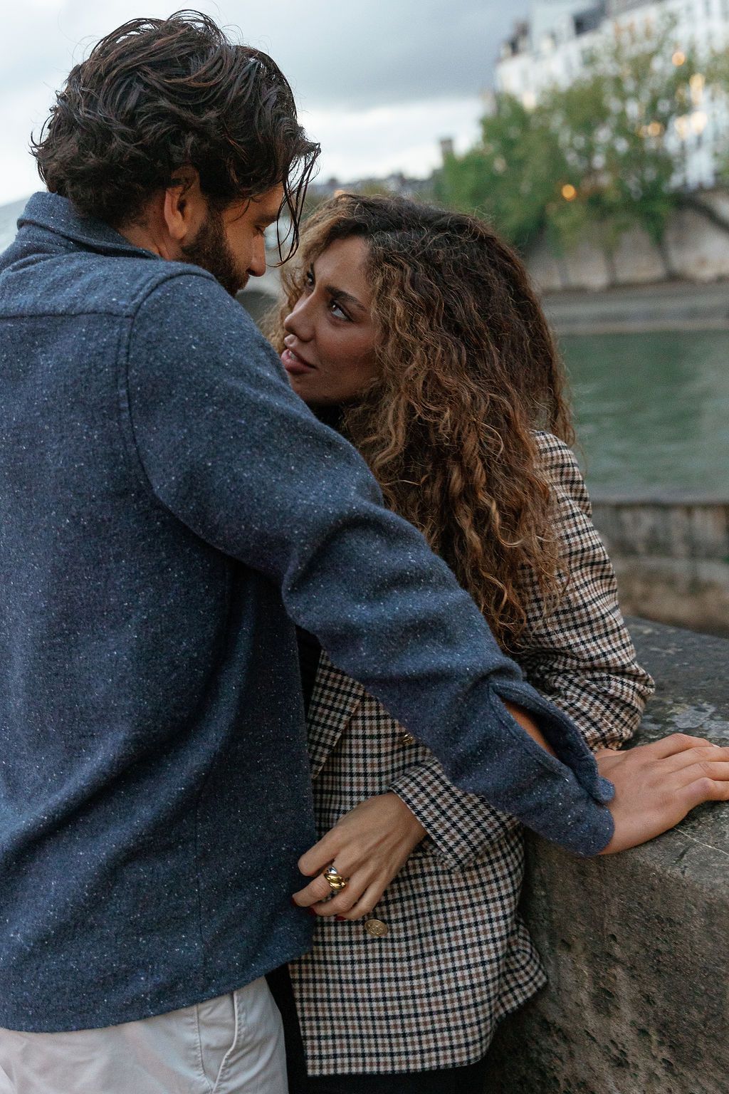 A couple embraces intimately along a stone wall beside a river on an overcast day in Paris, France.