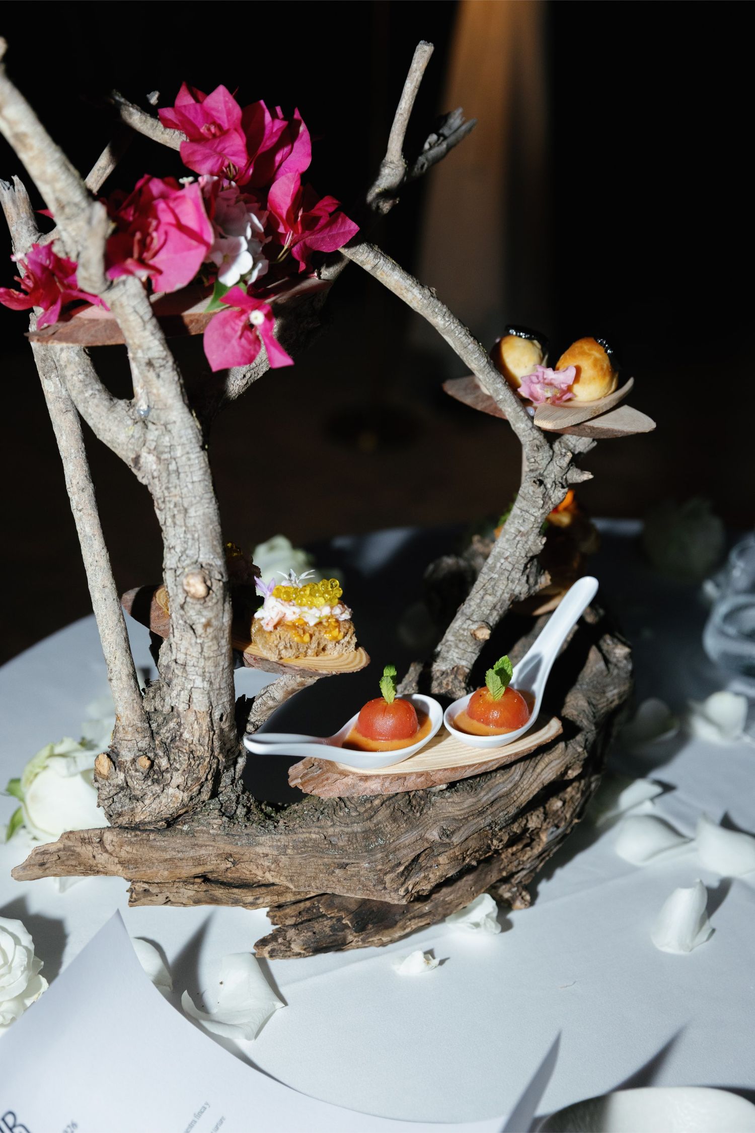 Artistic food presentation on driftwood with pink flowers, featuring elegant appetizers on white spoons and decorative branches.