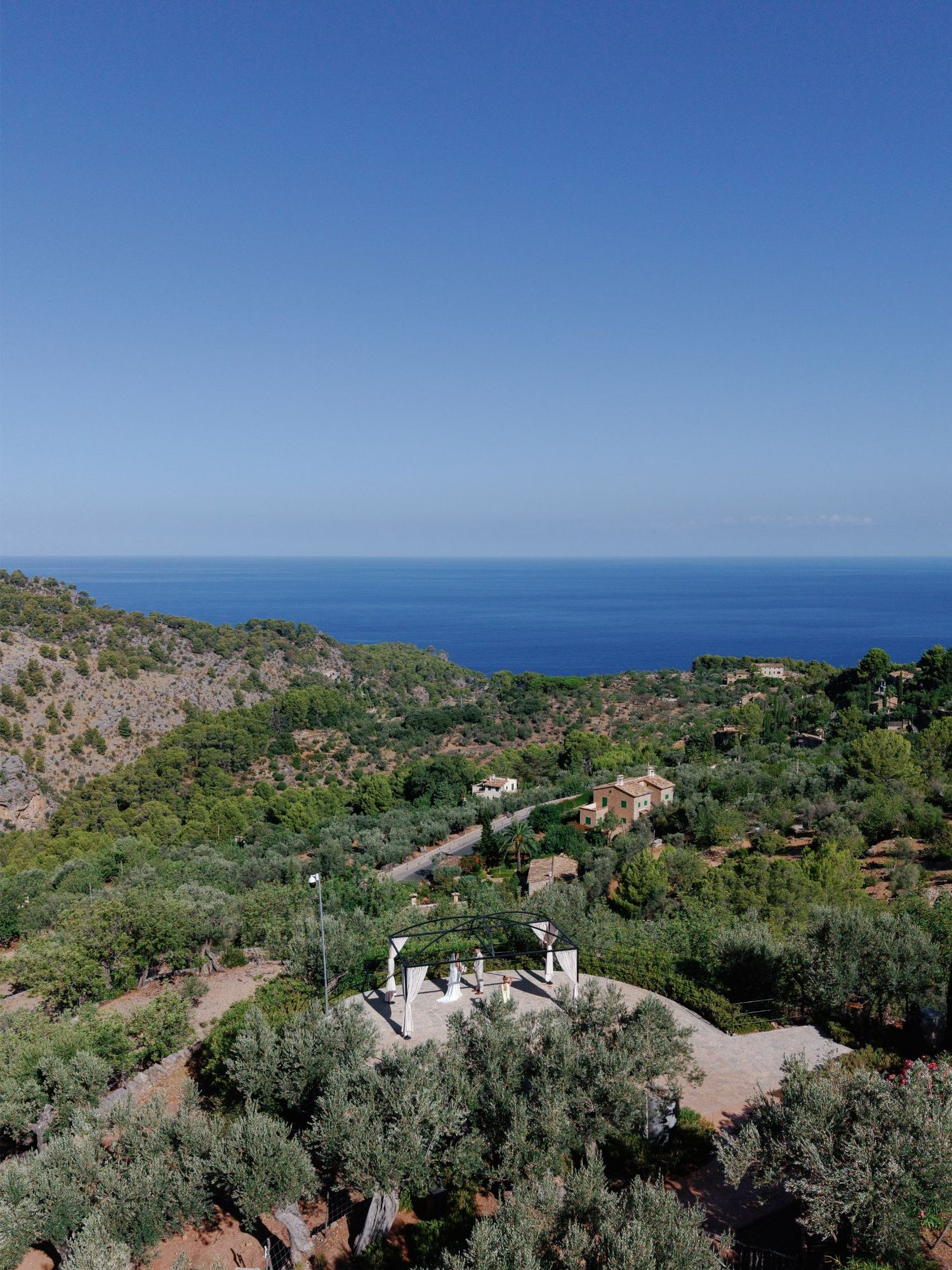 Aerial view of a Mediterranean hillside village surrounded by olive trees with stunning blue sea views under clear skies.