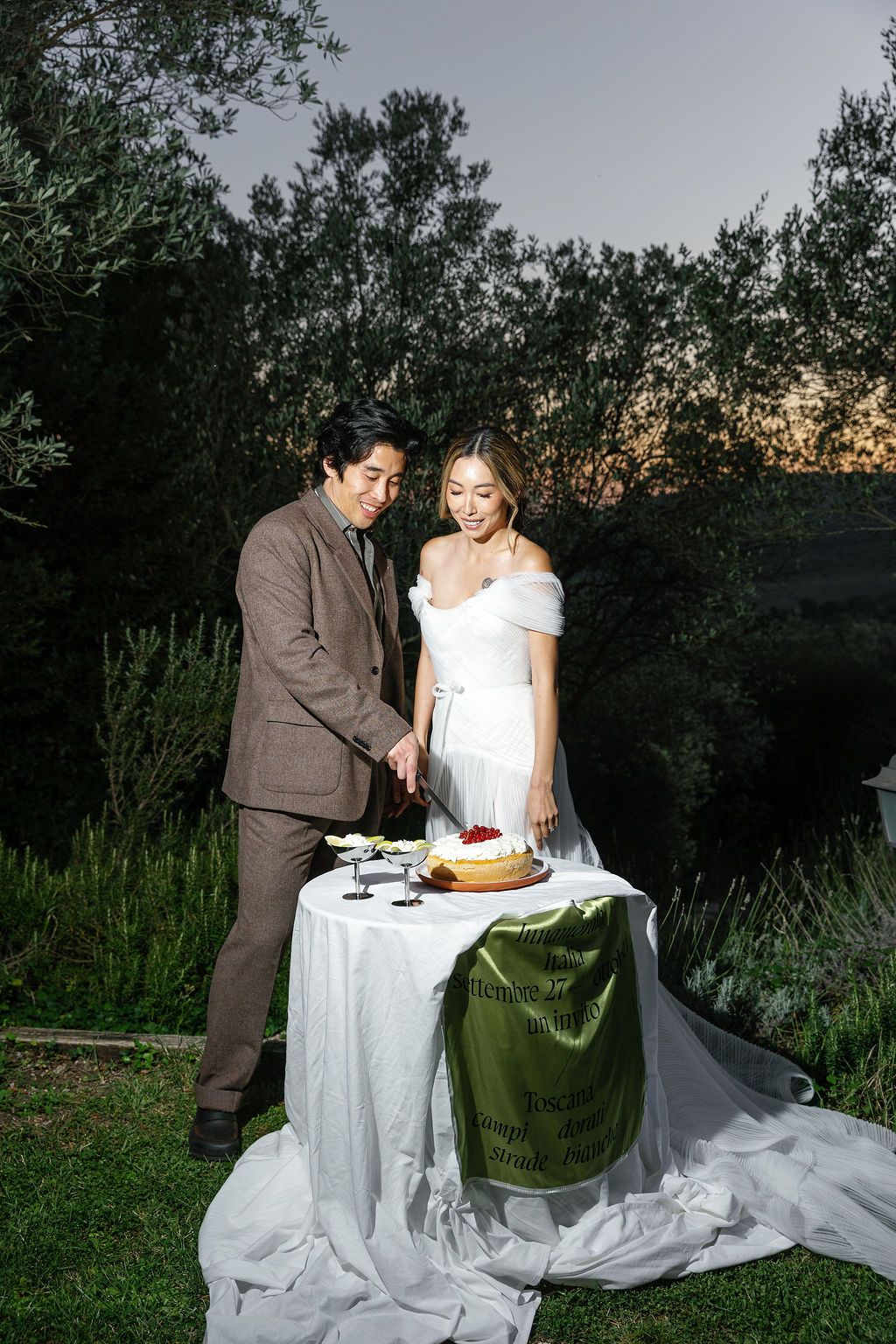 A couple cuts their wedding cake at an outdoor reception table as dusk settles in in Tuscany, Italy.