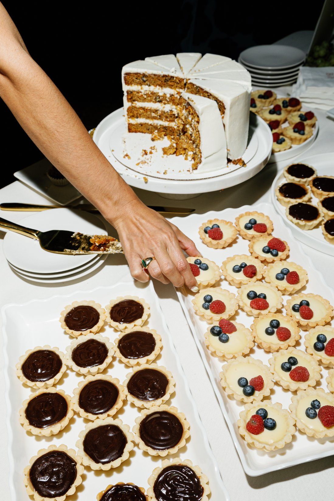 Hands arranging mini fruit tarts and chocolate desserts on white serving plates.