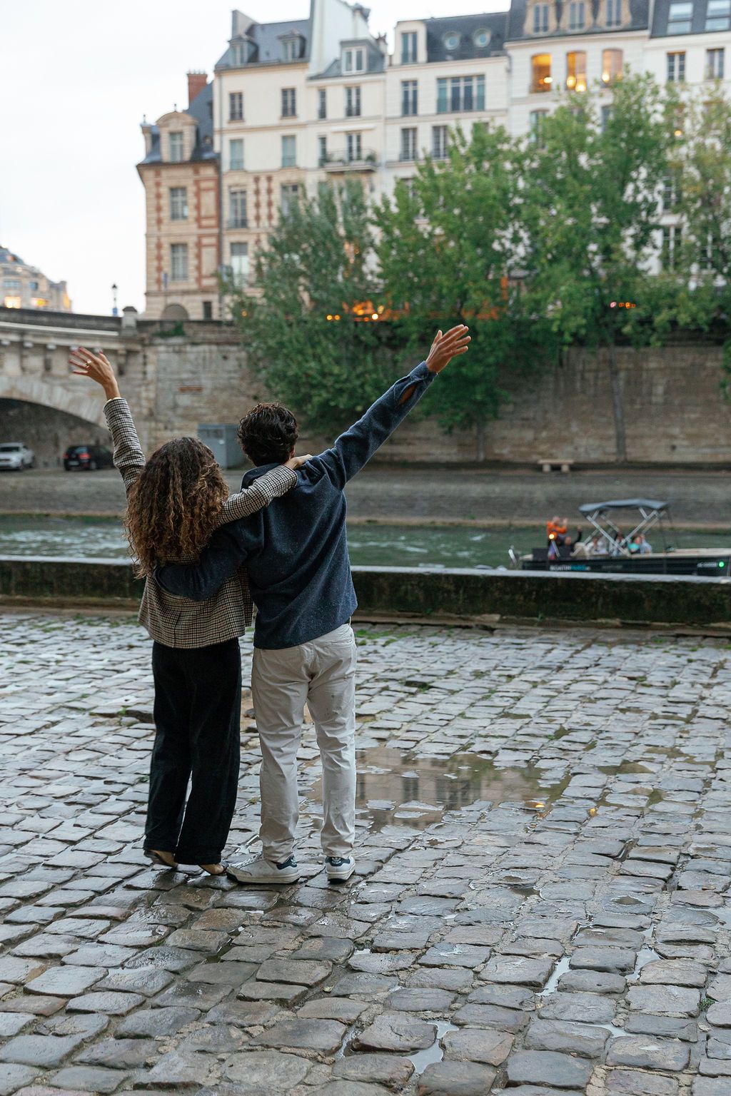 Two people stand together on a cobblestone path by a river, raising their arms in joyful celebration in Paris, France.