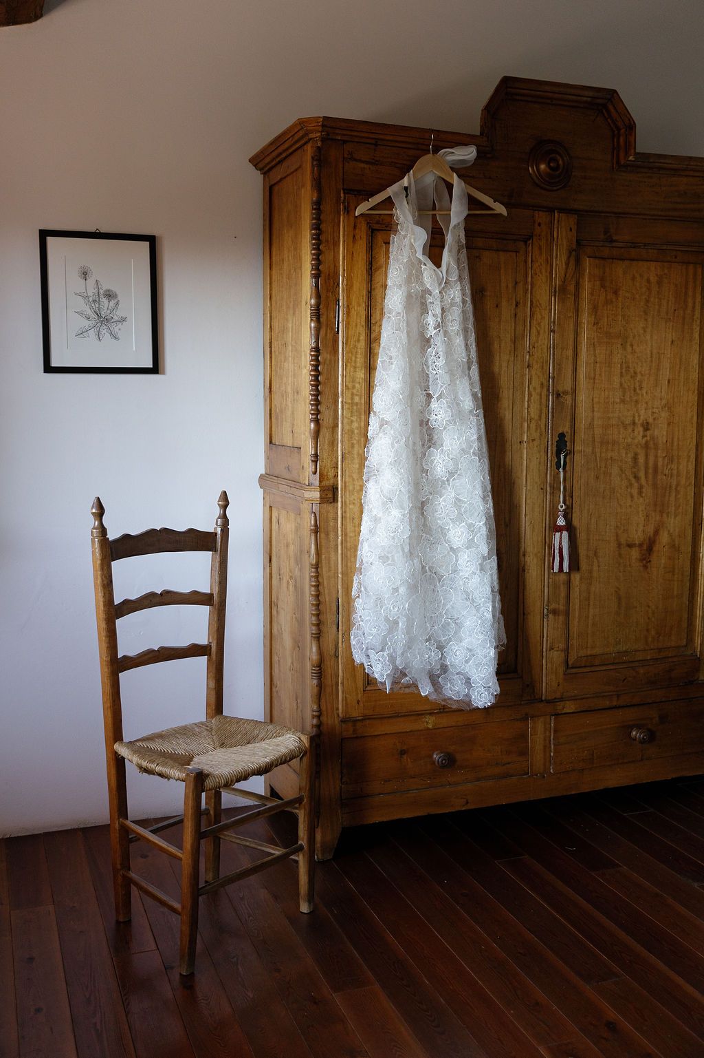 White lace wedding dress hanging on wooden armoire next to rustic chair in sunlit room in Tuscany, Italy.