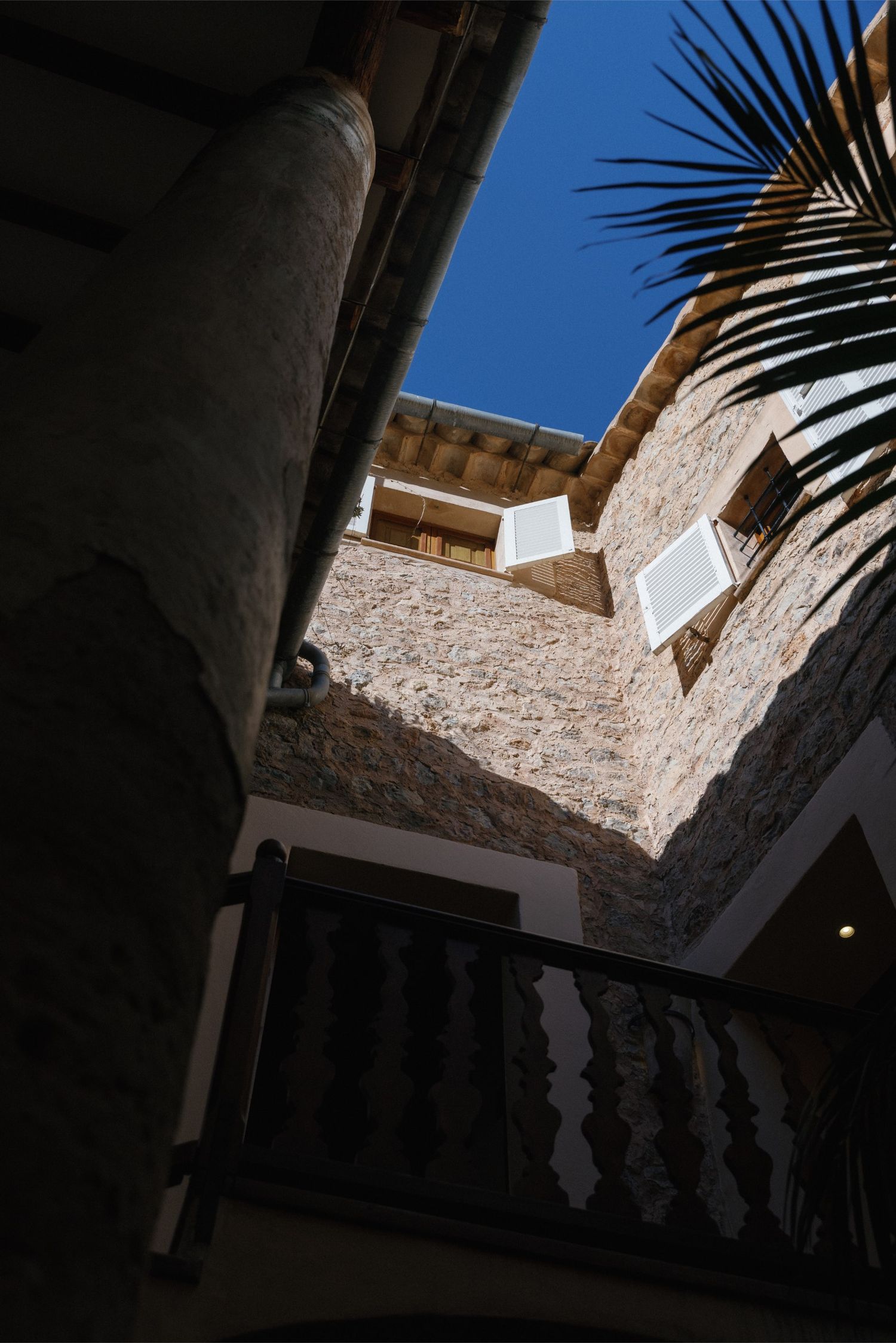 Upward view of a rustic stone building with white shuttered windows, ornate balcony railing, and palm tree against blue sky.