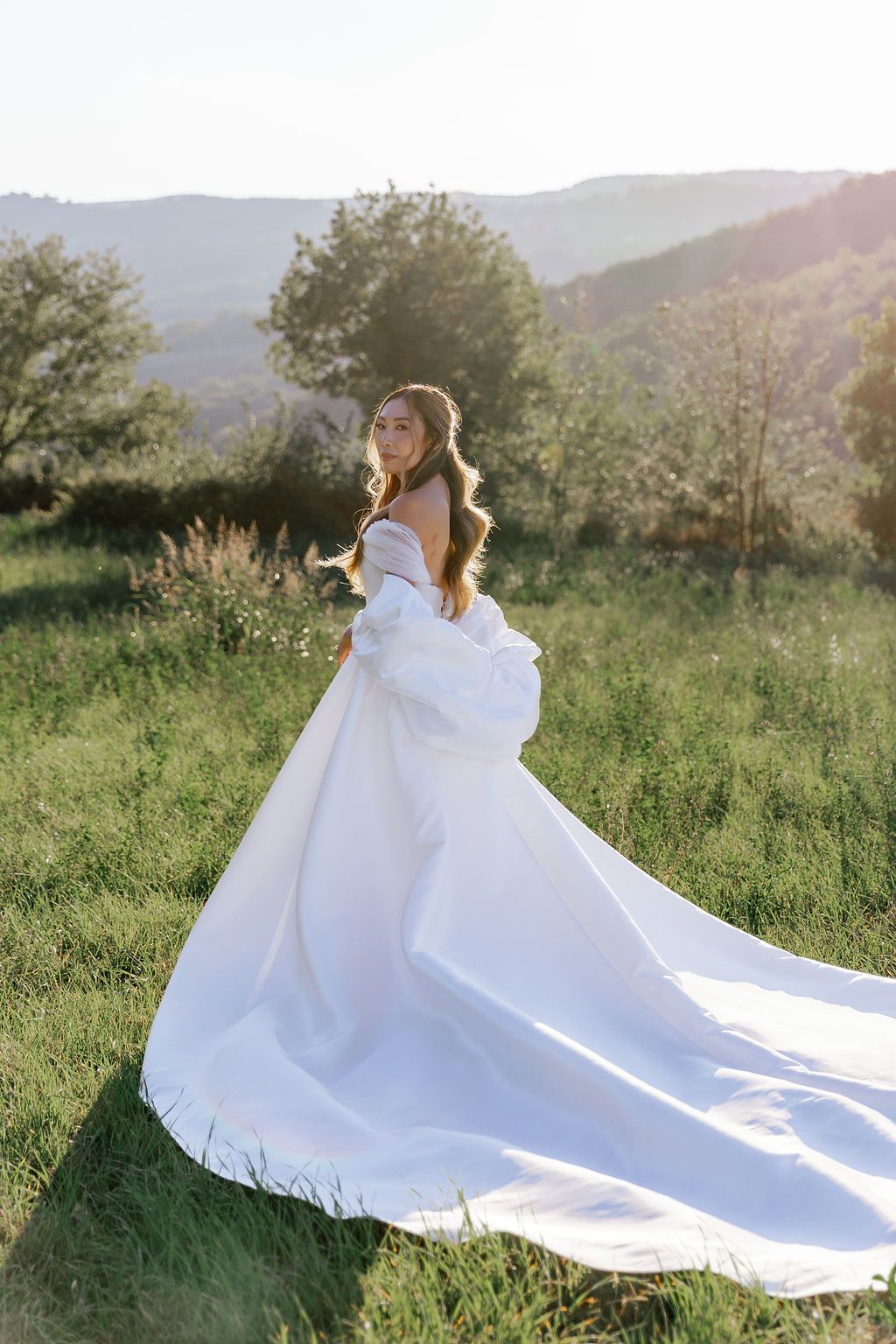 A flowing white wedding dress train spreads across a sunlit grassy hillside meadow with mountains in the background in Tuscany, Italy.