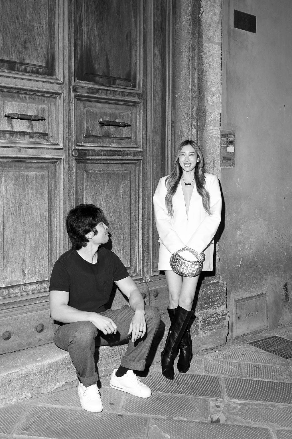 Black and white photo of two people sitting and standing on stone steps near a vintage wooden door in Tuscany, Italy.