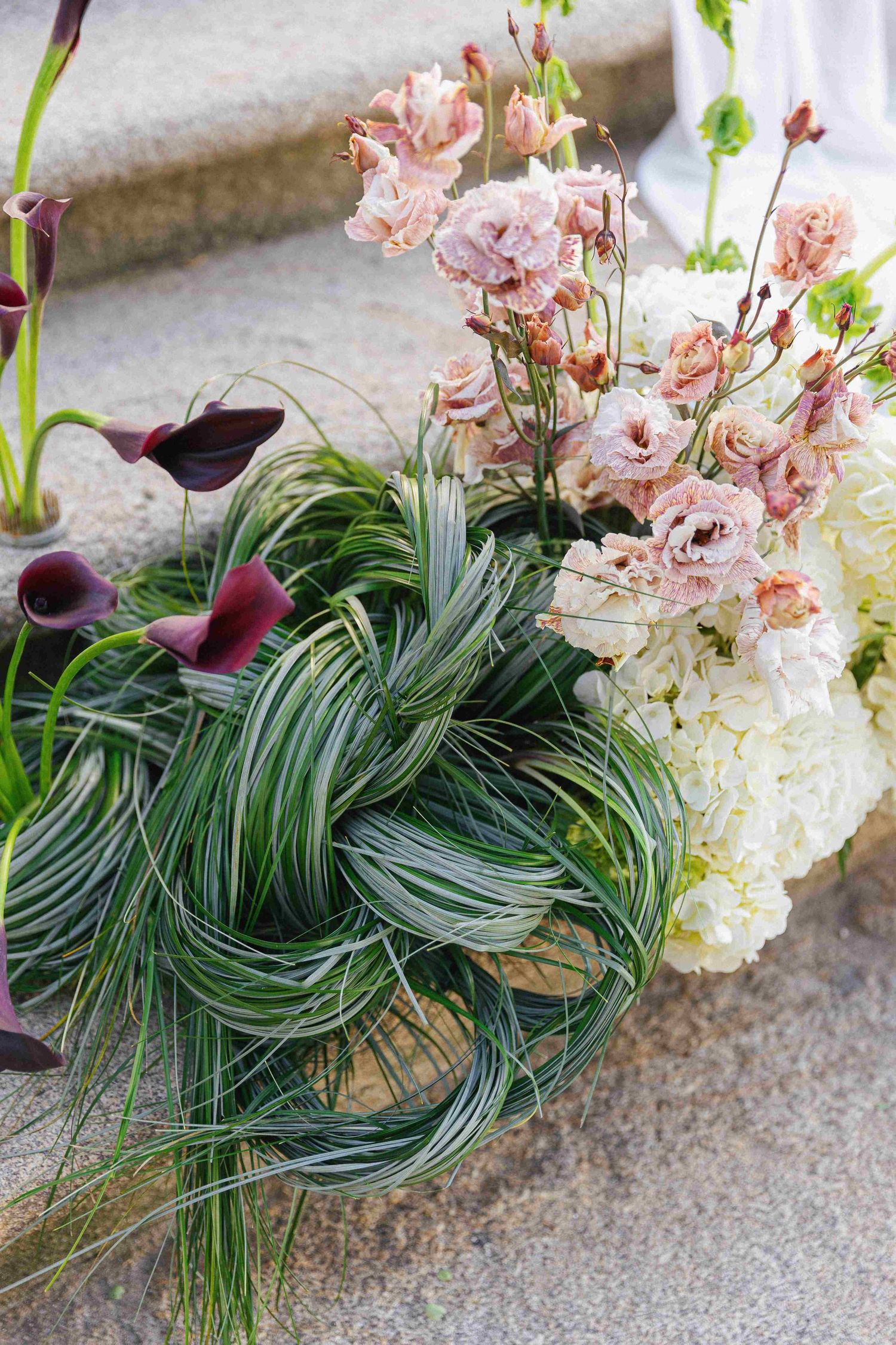 Elegant floral arrangement with white hydrangeas, pink orchids, dark calla lilies, and braided grass on stone steps.