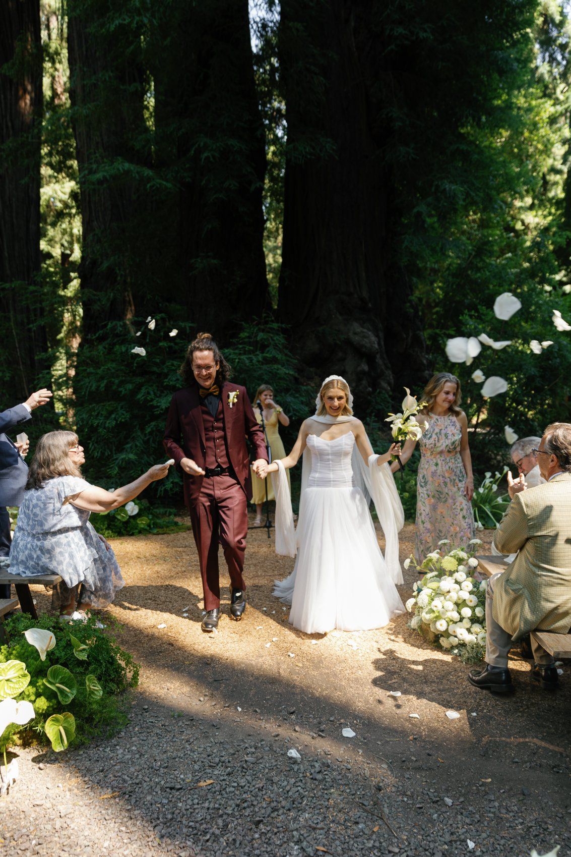 Wedding party members tossing white flower petals in a festive celebration on a forest path.