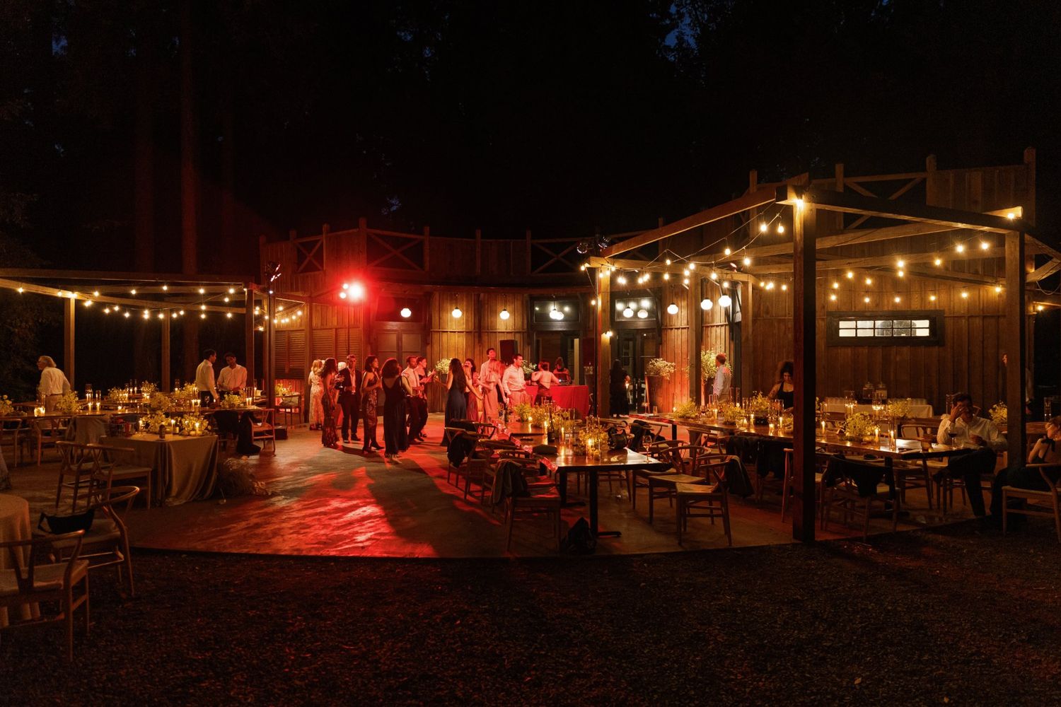 An outdoor wedding reception at night with string lights illuminating tables and guests under a covered pavilion.