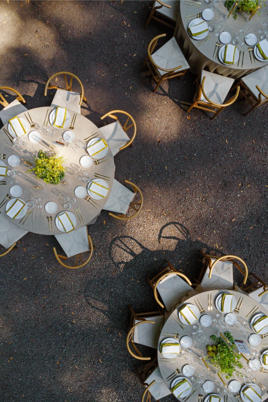 Overhead view of elegant round dining tables with white tablecloths and wooden chairs arranged on dark ground with dappled sunlight.